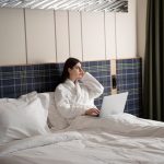 young-woman-working-her-laptop-while-sitting-bed-hotel-room_23-2149304021